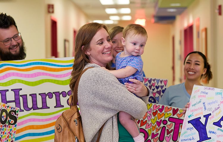 A smiling mother holding her child and Children's Mercy patient in the hall of the hospital. They are surrounded by people smiling and holding signs of encouragement.