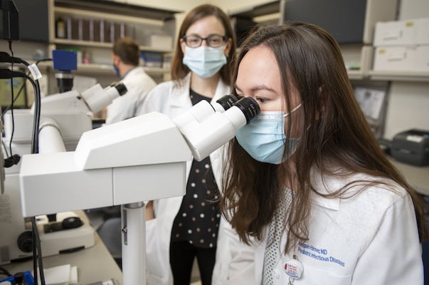 A masked young adult woman looks into a microscope as another female clinician looks on.