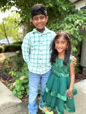 Young Indian boy in plaid shirt smiling with arm around younger sister in green dress.