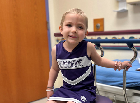 Children's Mercy patient, Elsie, is smiling while sitting on a stool in an exam room. She is wearing a K-State cheerleading uniform.
