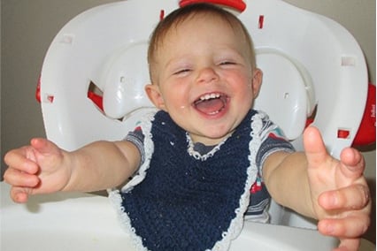 Dallin Mecham as a toddler sitting in a high chair, wearing a bib  and smiling with his arms outstretched.