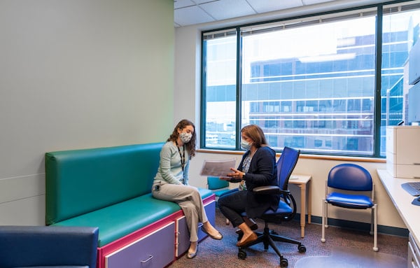 Two masked, female medical professionals talk together in an exam room setting.