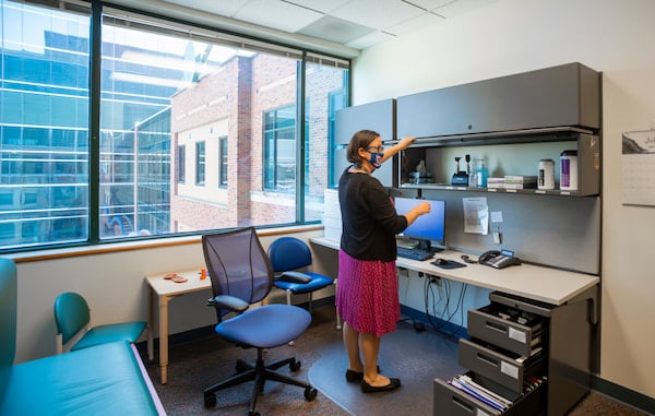 A masked, female medical professional opens a cabinet in an office.