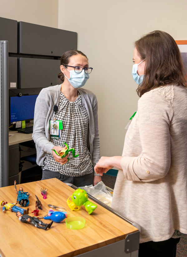 Two masked, female medical professionals sort through children's toys in an office environment.