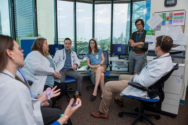 Several male and female clinicians, many in white coats, interact in an office setting.