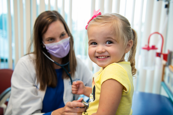 A young girl smiles as a masked, female clinician uses a stethoscope on her torso.