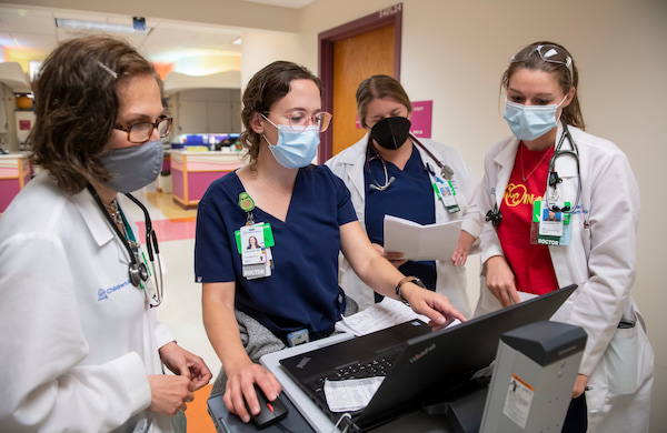 Four masked, female physicians look at a laptop together in a hospital hallway.