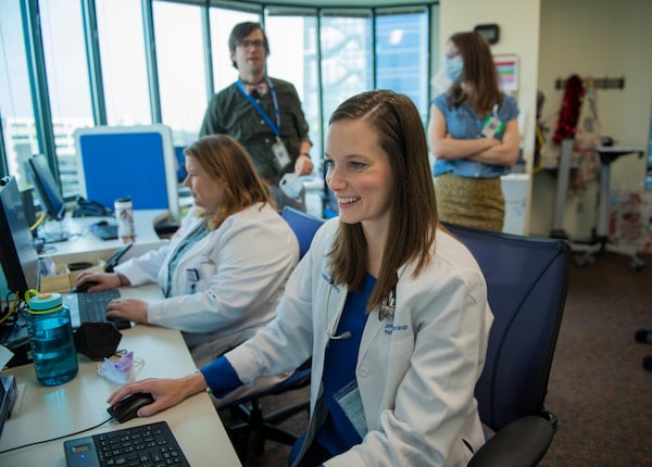 A smiling, female physician looks at a computer monitor while three other clinicians interact behind her in an office setting.