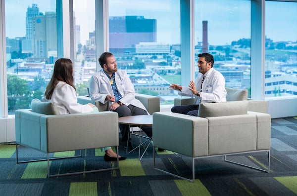 Three physicians, two male and one female, sit and chat next to a window overlooking Kansas City.