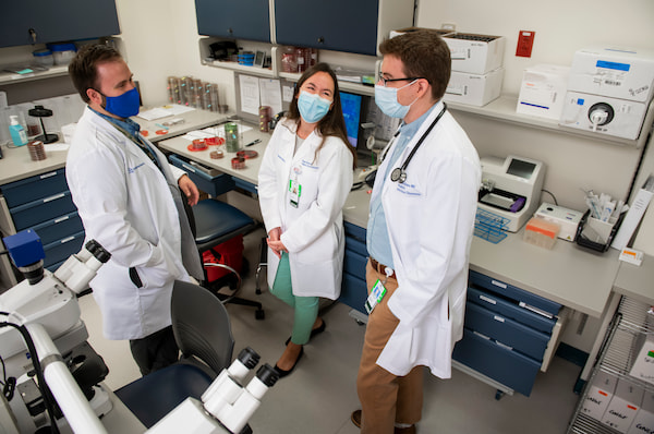 Three physicians in white coats and masks chat in a laboratory setting.