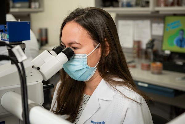 A masked female physician looks into a microscope.