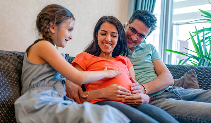 Young child touches mom's pregnant belly while sitting next to her and dad on the couch.