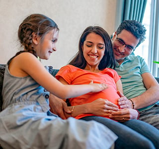 Young child touches mom's pregnant belly while sitting next to her and dad on the couch.