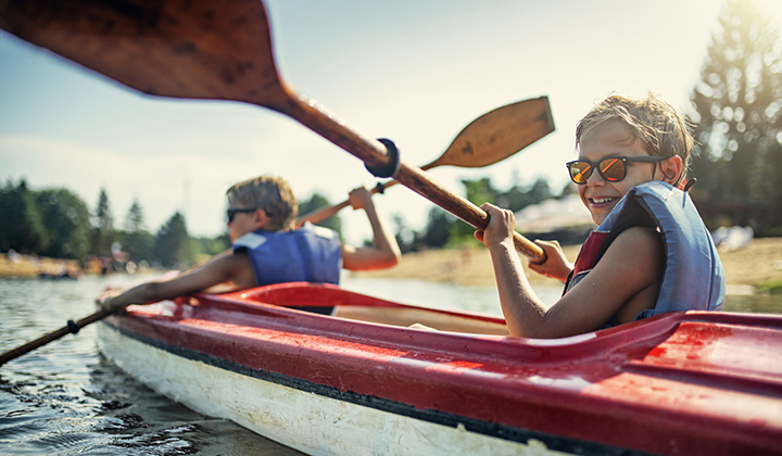 Two young boys in a canoe and wearing life jackets. They are using rowing paddles on a lake and smiling.