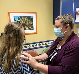 Young patient with back to camera gets hear heart listened to by a doctor in a clinic room.