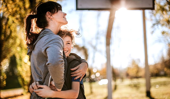 Mom hugs her preteen son. They are wearing sports clothes and near a basketball goal outside on a sunny day.