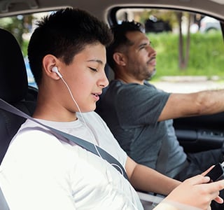 Teen boy sits in the passenger seat of the car with his dad listening to a phone with headphones.