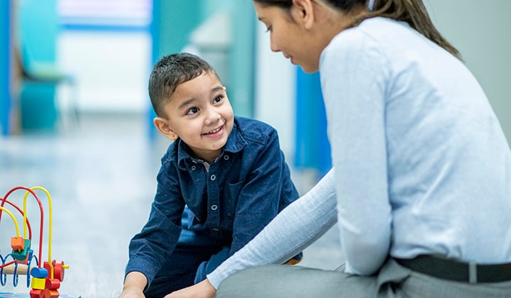 Young boy wearing a dark blue shirt sits on the floor next to a woman in a light blue shirt. They are playing with a toy on the ground.