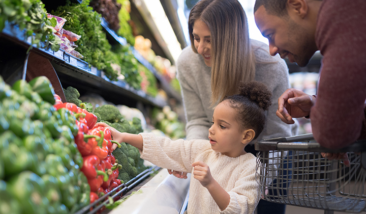 Young child reaching for red bell pepper in grocery store with parents behind her.