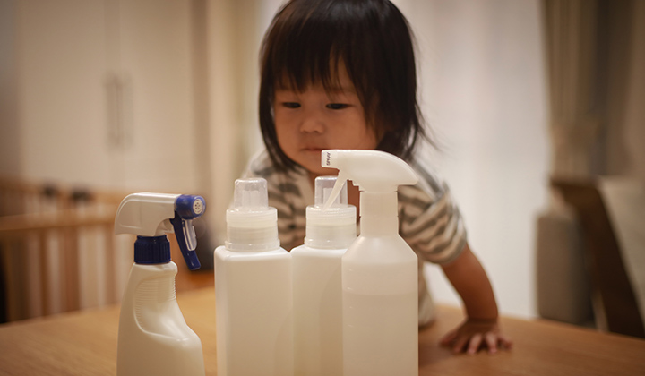 Toddler girl wearing a grey striped shirt leans over a table to look at four white cleaning products.