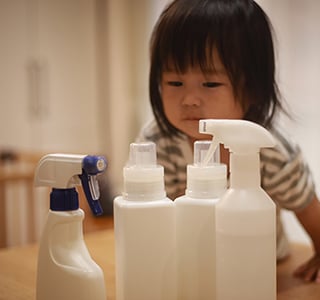 Toddler girl wearing a grey striped shirt leans over a table to look at four white cleaning products.