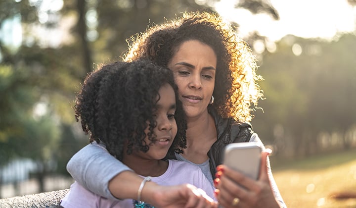 Mom hugs her preteen daughter and looks at a cell phone.