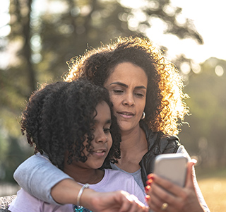 Mom hugs her preteen daughter and looks at a cell phone.