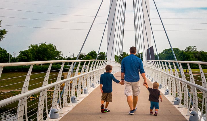 Brandon Billinger holds hands with his two young sons walking away as the cross a walking bridge on a sunny day.