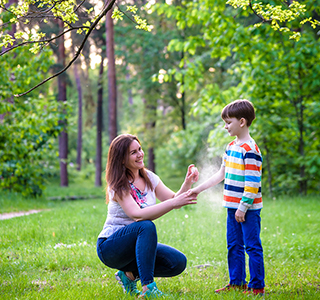 Woman sprays bug repellant on young child while outside in a woody area.
