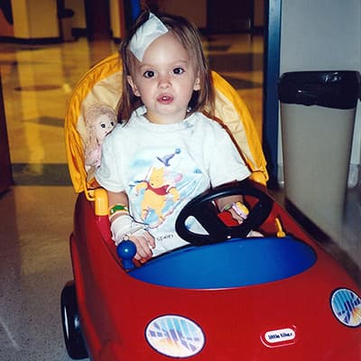 Toddler girl wearing a Winnie the Pooh shirt with a large bandage on the top of her head. She is driving a play car in a hospital.