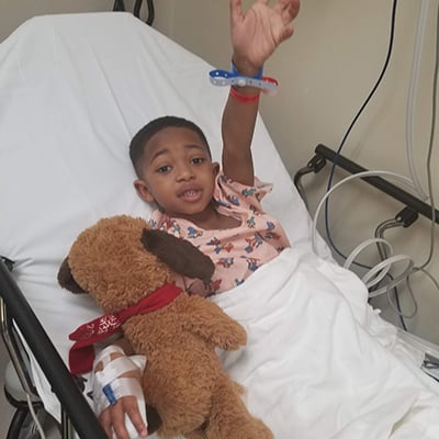 A toddler in a hospital gown waves at the camera from a hospital gurney. He has an IV taped to one hand and hospital bracelets around the other wrist. He’s holding a stuffed animal.