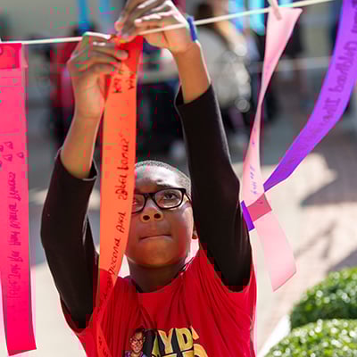 A boy in glasses concentrates as he attaches an orange ribbon with writing 	on it to a string. There are more people and ribbons out-of-focus behind him. He is wearing a t-shirt with a cartoon version of himself and lettering that reads “Ayden: 	Kid Hero.”