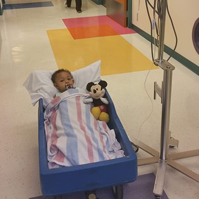 A baby looks at the camera from a blue hospital wagon. He’s covered by a blanket and has a Mickey Mouse doll beside him. There is an IV next to the wagon and a hospital hallway in the background.