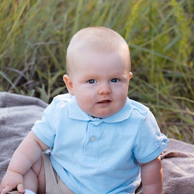 A baby with full cheeks and blond hair sits on a blanket in a field of grass. He is wearing a light blue polo shirt and khaki shorts.