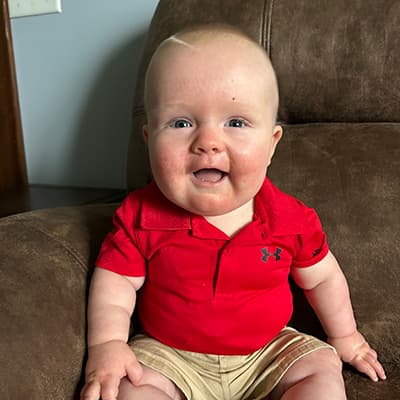 A blond, round-cheeked baby boy smiles at the camera. He’s wearing a red polo shirt and sitting in a big brown chair. 