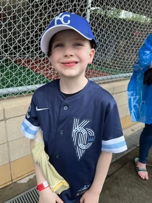 A young boy smiles while wearing a Kansas City Royals jersey and an oversized baseball cap.