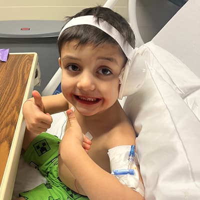 A little boy with brown hair sits up in a hospital bed. He has a protective bandage over his left ear and around his head and an IV connection in his arm. He is wearing Minecraft shorts, smiling and giving 2 thumbs up to the camera.