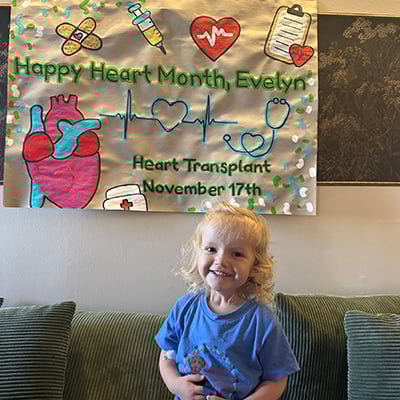A toddler with blonde hair and a big smile stands on a green couch under a big sign that says “Happy Heart Month, Evelyn! Heart Transplant November 17th.” She’s wearing a blue shirt, and she has tape and a cotton ball on one inner arm, as if she just had blood drawn.  