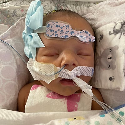 Close up of a small infant with medical tubes coming out of her nose and mouth. She has a medical monitor on her forehead beside a blue bow. She’s covered in soft blankets.  