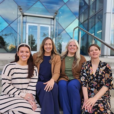 Four women sit on the steps in front of a glass building. They are all smiling and are dressed in office attire in autumnal colors. 