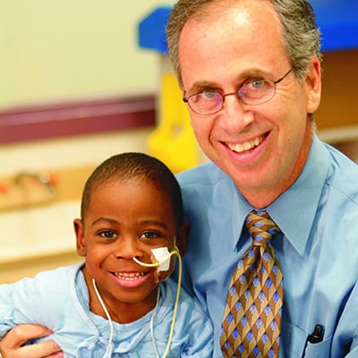 An adult and a young child sit together closely, smiling, with the child wearing medical tubing on the face.
