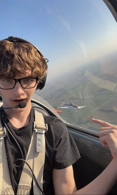 teen boy grins and points out the window of the small airplane he is flying. Through the window, you can see another small aircraft