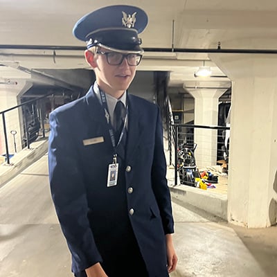 A tall teen boy poses in a parking garage. He is wearing a navy blue formal military uniform, including a suit jacket, creased pants and a white dress shirt and blue tie. He is wearing a blue cap with a black brim and a gold eagle insignia.