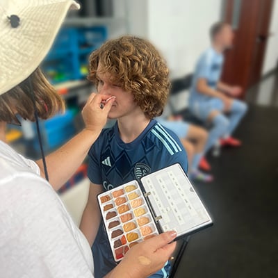 A makeup artist applies makeup from a palette of various skin tones to a teen boy wearing a soccer uniform.