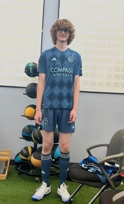 A tall teen boy with very curly hair stands in a locker room. He is wearing a blue Sporting KC uniform and smiling.