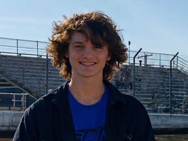 A teen boy with very curly hair stands in a locker room. He is wearing a blue Sporting KC uniform and smiling.