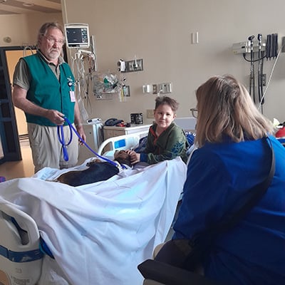Jaxson sits in a hospital bed interacting with a therapy animal while two adults visit in the patient room.