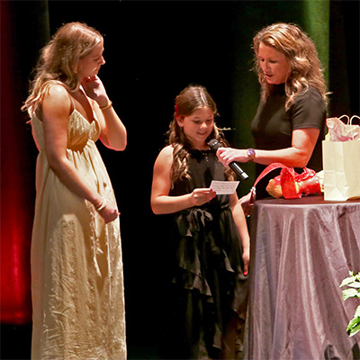 Three people stand on a stage. Jenna reads from a note card while another person holds a microphone. Liv stands nearby wearing a light yellow dress. A table with gifts and decorations is positioned on the stage.