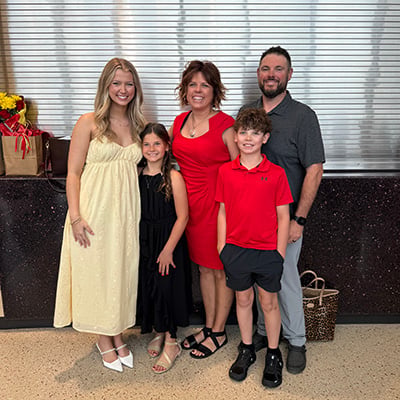Five people pose for a photo indoors following a ceremony. Jenna stands near the center with family members and Liv, who is wearing a light yellow dress. Gift bags are visible nearby.