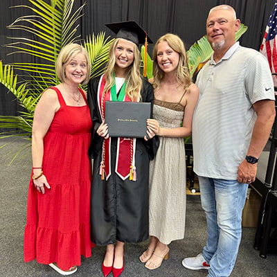 Four people pose together at a graduation ceremony. One person wears a graduation cap and gown and holds a diploma cover. Indoor plants and event draping are visible in the background.